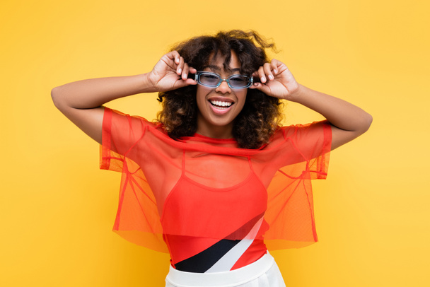happy african american woman in trendy summer clothes adjusting sunglasses isolated on yellow - Foto, Imagen