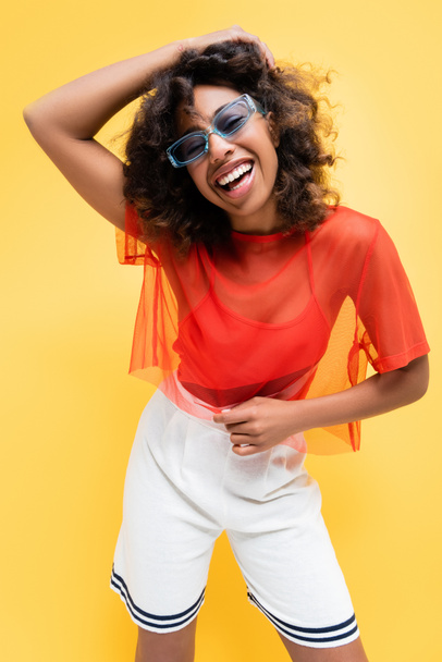 excited african american woman in white shorts and purple sunglasses laughing isolated on yellow - Фото, зображення