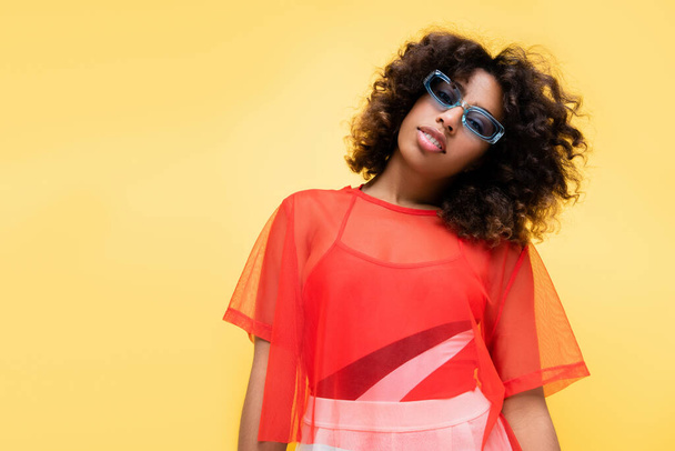 african american woman in stylish summer outfit and blue sunglasses isolated on yellow - 写真・画像