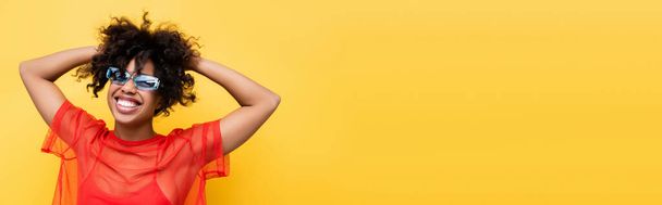 radosna Afroamerykanka w czerwonej bluzce i niebieskich okularach, pozująca na żółtym, sztandarze - Zdjęcie, obraz