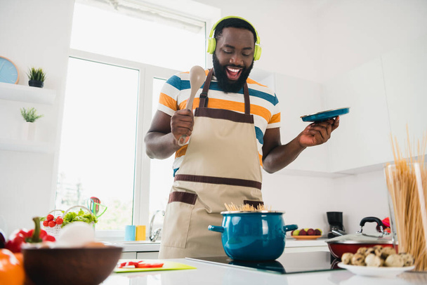 Portrait of funny cheerful handsome guy enjoy preparing delicious pasta listen music mp3 kitchen indoors - Φωτογραφία, εικόνα
