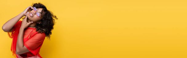 стильная африканская американка в солнечных очках касаясь подбородка изолированы на желтый, баннер - Фото, изображение