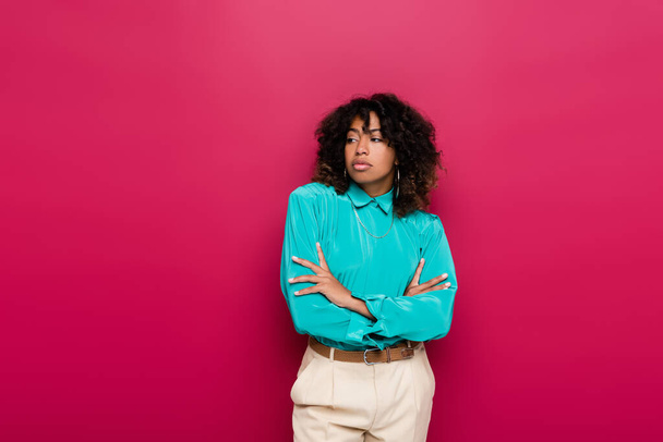 mujer afroamericana con el pelo ondulado de pie con los brazos cruzados y mirando hacia otro lado aislado en rosa - Foto, Imagen