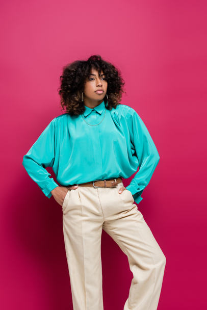 african american woman in turquoise blouse posing with hands in pockets of trousers isolated on pink - Фото, изображение