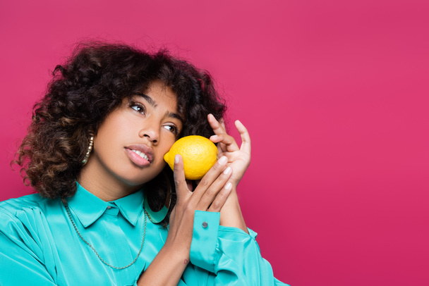 jolie femme américaine africaine regardant loin tout en posant avec citron entier isolé sur rose - Photo, image