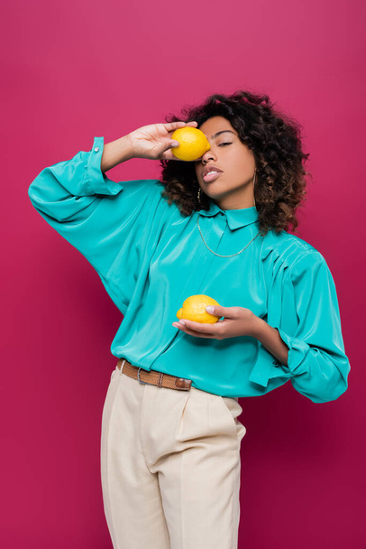 african american woman in turquoise blouse obscuring face with whole lemon isolated on pink - Foto, afbeelding