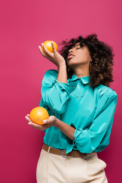 curly african american woman in stylish blouse posing with oranges isolated on pink - 写真・画像