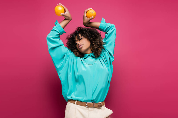 african american woman in turquoise blouse posing with oranges in raised hands  isolated on pink - Photo, Image