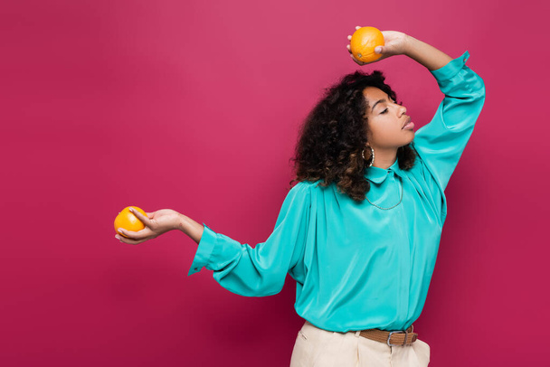 femme afro-américaine à la mode posant les yeux fermés et des oranges mûres isolées sur rose - Photo, image