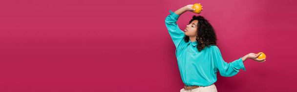 stylish african american woman in blue blouse posing with oranges isolated on pink, banner - Photo, Image