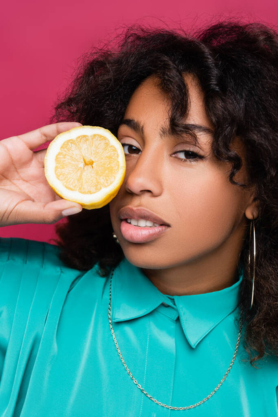 brunette african american woman holding half lemon near face isolated on pink - Фото, изображение