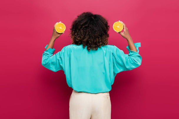visão traseira da mulher americana africana em blusa azul segurando metades de laranja isolado em rosa - Foto, Imagem