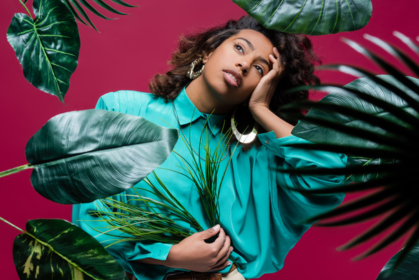 african american woman in turquoise blouse holding hand near face while posing with tropical leaves isolated on pink - Valokuva, kuva