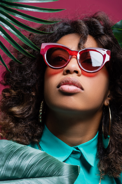 Afroamerykanka w modnych okularach przeciwsłonecznych i kolczykach odwracając wzrok od zielonych liści odizolowanych na różowo - Zdjęcie, obraz