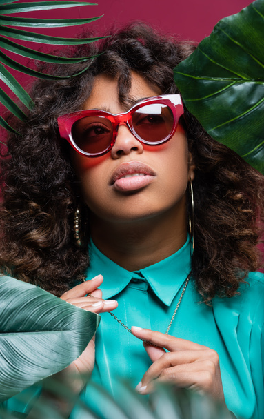 african american woman in stylish sunglasses touching necklace near exotic leaves isolated on burgundy - Foto, Imagem