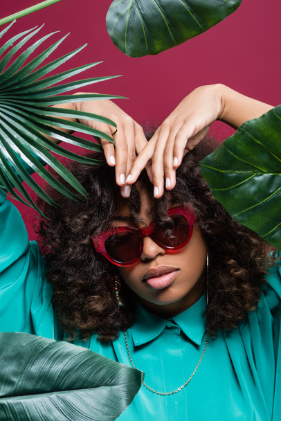 african american woman in sunglasses posing with hands above head near leaves isolated on pink - Fotografie, Obrázek