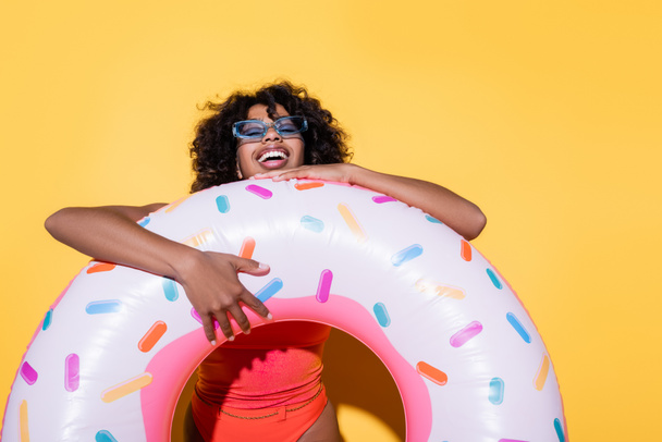 excited african american woman in trendy sunglasses laughing near swim ring on yellow background - Photo, Image