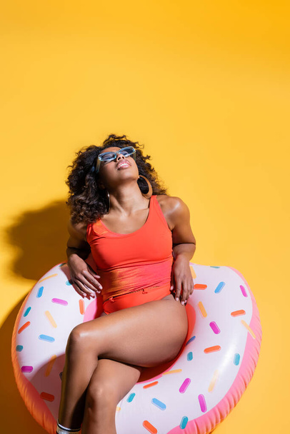 top view of african american woman in swimsuit relaxing on inflatable ring on yellow background - 写真・画像