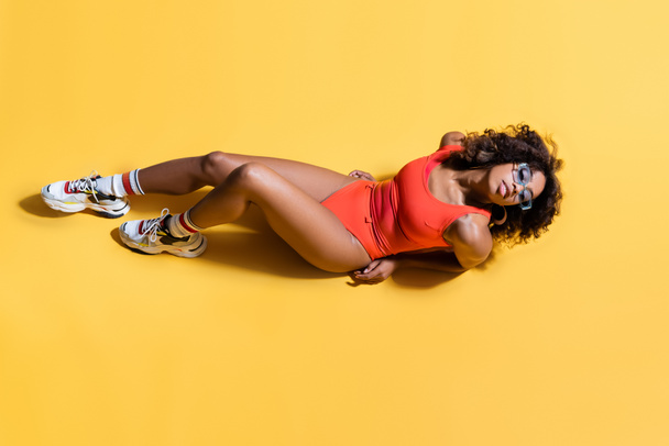top view of sensual african american woman in red swimsuit, sunglasses and sneakers on yellow background - Zdjęcie, obraz