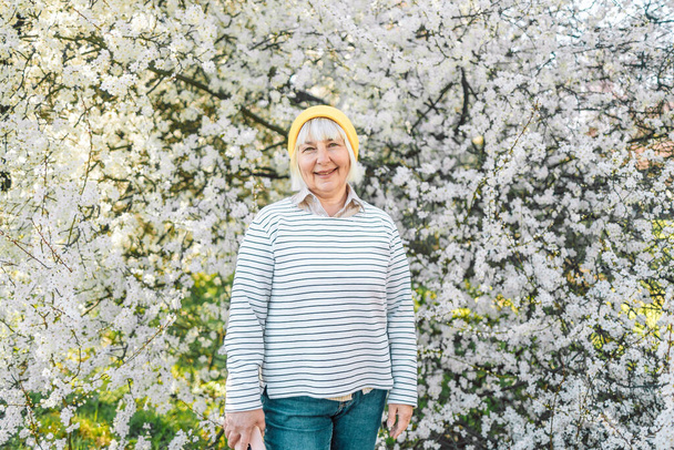 Porträt einer glücklichen schönen blondhaarigen Seniorin in stylischer Kleidung und gelber Mütze, die im Frühling allein im Park in die Kamera lächelt - Foto, Bild