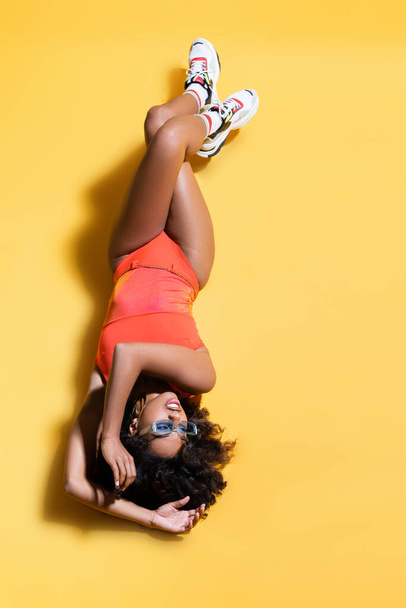 top view of african american woman in swimsuit and sneakers lying with crossed legs on yellow background - Fotoğraf, Görsel
