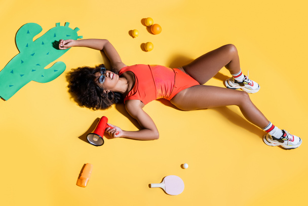 stylish african american woman lying with megaphone near paper cut cactus, fruits and sunscreen on yellow background - Photo, Image
