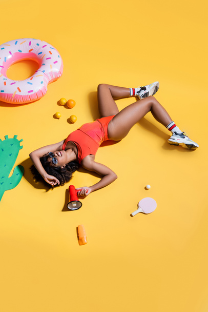 donna afroamericana in sneakers e costume da bagno sdraiata con megafono vicino ai frutti e anello nuoto su sfondo giallo - Foto, immagini