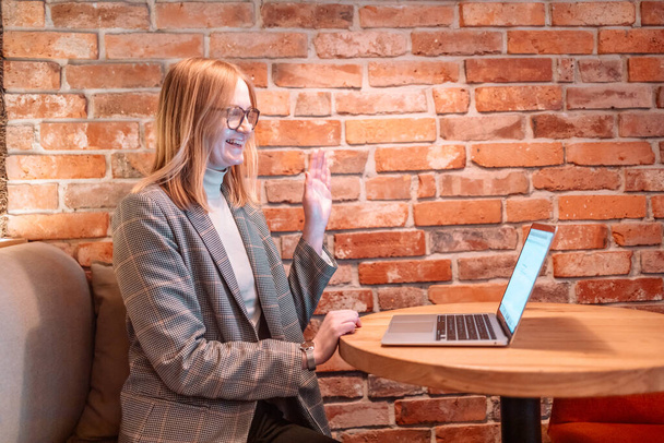 Позитивная студентка разговаривает, машет рукой, используя ноутбук компьютерной видеоконференции, сидя за столом в кафе на открытом воздухе.  - Фото, изображение