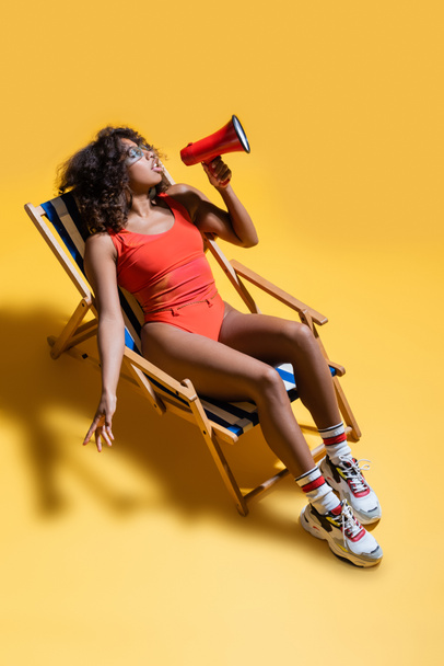 afrikkalainen amerikkalainen nainen trendikkäissä rantavaatteissa istuu kansituolissa ja puhuu megafonilla keltaisella taustalla - Valokuva, kuva