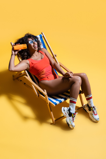 happy african american woman in swimwear and sneakers resting in deck chair and holding sunscreen lotion on yellow - Фото, изображение