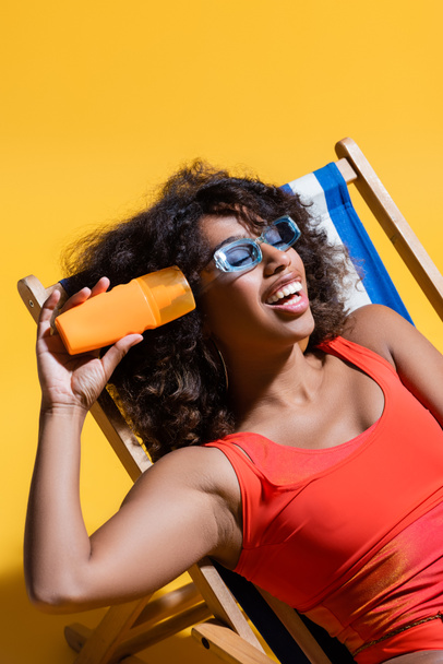 alegre afroamericana mujer en gafas de sol de moda sentado en cubierta silla con protector solar sobre fondo amarillo - Foto, Imagen