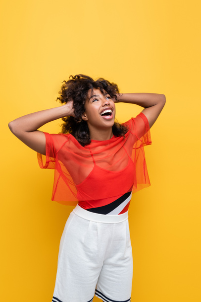 amazed african american woman in summer outfit touching wavy hair and looking up isolated on yellow - Фото, изображение