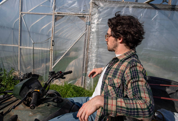 Mladý zemědělec ve flanelové košili sedí na terénní vozidlo mimo skleník - Fotografie, Obrázek