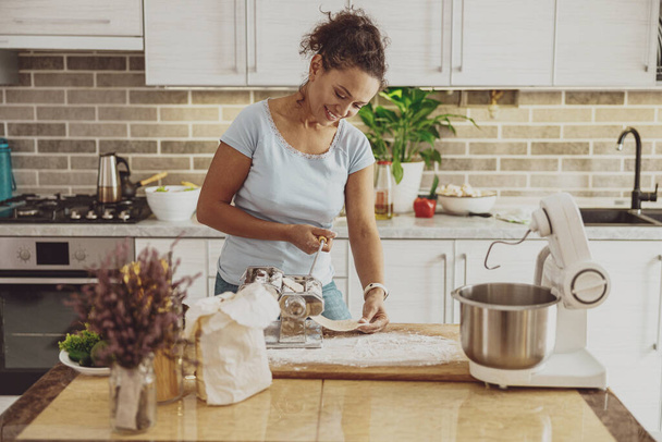 台所の笑顔の女性は、生地ミキサーと麺カッターを使用して麺を準備します - 写真・画像