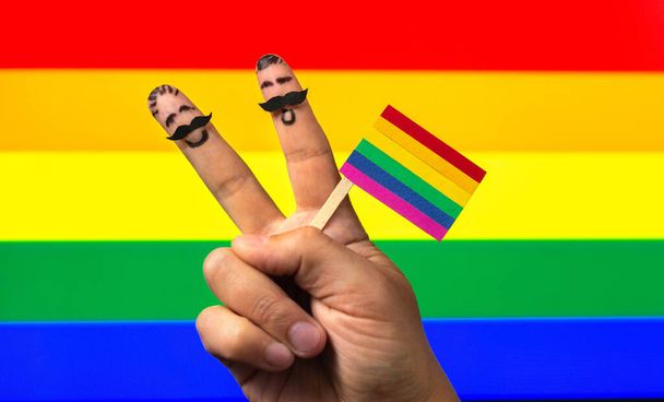 las manos con dos dedos hombres sosteniendo la bandera del arco iris, signo de paz. Día internacional contra la homofobia, la transfobia y la bifobia - Foto, imagen
