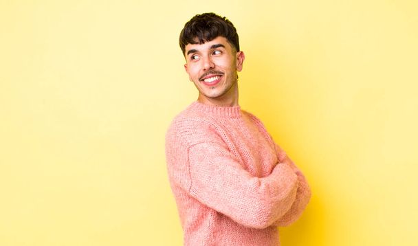 young adult hispanic man smiling gleefully, feeling happy, satisfied and relaxed, with crossed arms and looking to the side - 写真・画像
