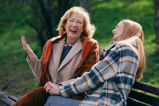 Бабуся сміється і розважається зі своєю підлітковою онукою, сидячи на лавці парку
. - Фото, зображення