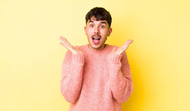 young adult hispanic man looking happy and excited, shocked with an unexpected surprise with both hands open next to face - Foto, Imagen