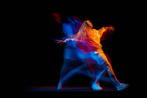 Young woman dancing hip-hop in sportive style clothes isolated on dark background at dance hall in mixed neon light. Youth culture, hip-hop, movement - 写真・画像