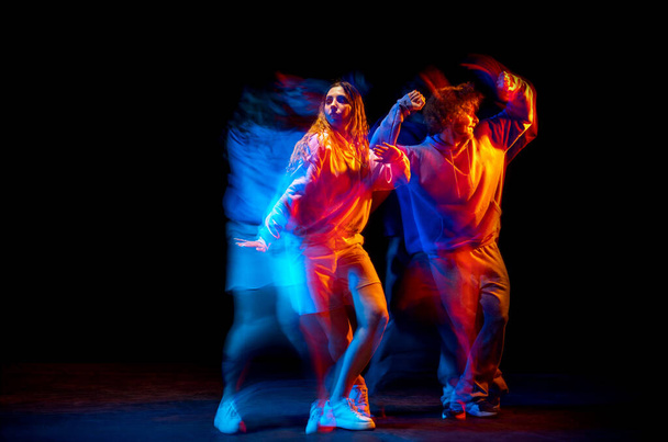 Young man and woman dancing hip-hop in sportive style clothes on dark background at dance hall in mixed neon light. Youth culture, hip-hop, movement - Zdjęcie, obraz
