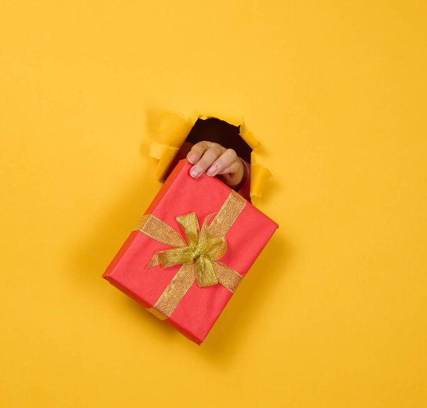 female hand holds a box with a gift on a yellow background, part of the body sticks out of a torn hole in a paper background. Congratulation, holiday surprise - Foto, afbeelding