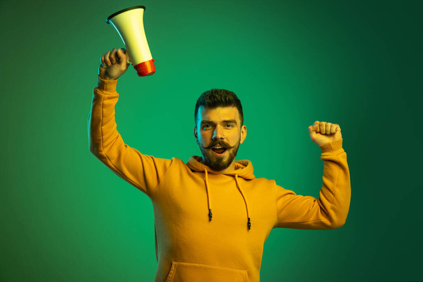 Portrait of young man in casual cloth shouting in megaphone isolated over green studio background. Party time - Foto, Imagem