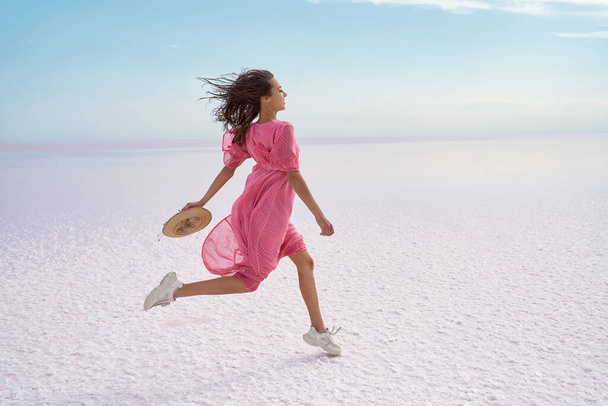Running carefree woman on salt flats on pink lake. Brunette model in pink adorable flowing dress - Fotoğraf, Görsel