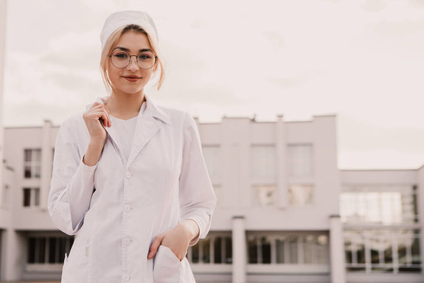 Junge Krankenschwester steht vor der Krankenstation des Krankenhauses. Wunderschöne Ärztin in weißem Arztkittel und Mütze mit Brille posiert lächelnd in die Kamera. - Foto, Bild