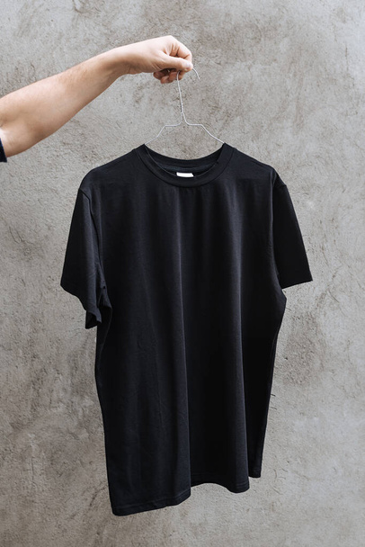 Front view of a black t-shirt hanging on a clothes hanger on a background of textured concrete wall. The male hand holds it as a presentation. Design and layout of men's t-shirt for printing. - Fotó, kép