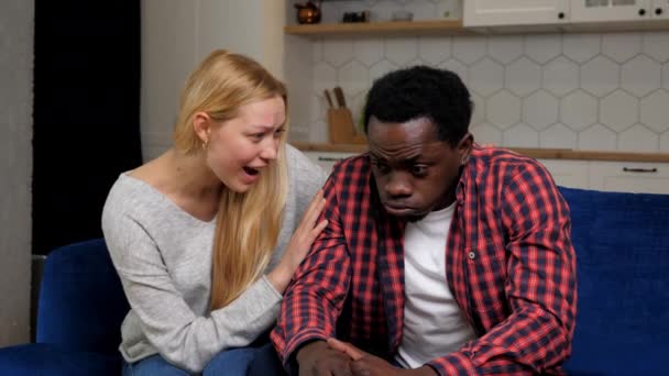 Smiling caucasian woman calms African American upset man sitting on couch at home kitchen. Female motivates male by giving motivational speech. Multiethnic family couple talking about relationships - Felvétel, videó