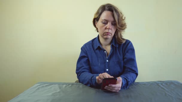 Egy negyven éves nő kék farmeringben ül egy asztalnál, kezében egy mobiltelefonnal. Lassan mozgó portré. Felnőtt szőke nő - Felvétel, videó