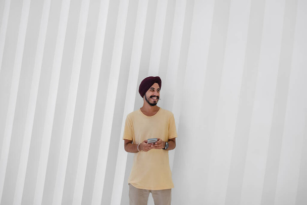 Portrait d'un homme heureux utilisant un téléphone portable au centre commercial. Joyeux homme indien souriant tapant un message texte sur son smartphone tout en se tenant devant un mur géométrique blanc et en regardant loin. - Photo, image