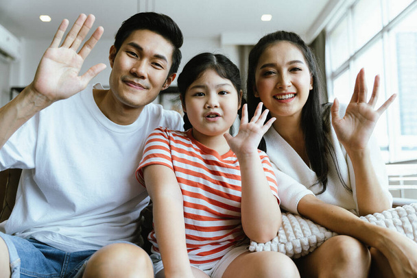 Mini happy family parent and cute girl kid looking and raised hand up to camera to greeting with the other side of online network. Online greeting to keep social distancing. - Foto, afbeelding