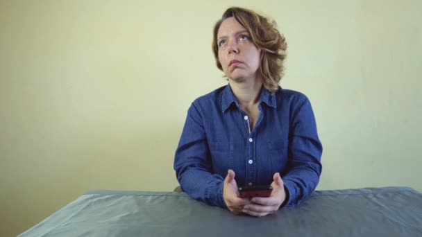 Een 40-jarige vrouw in een denim shirt zit aan een grijze tafel. Een volwassen vrouw denkt en houdt een mobiele telefoon in haar handen. Langzame beweging 4k - Video